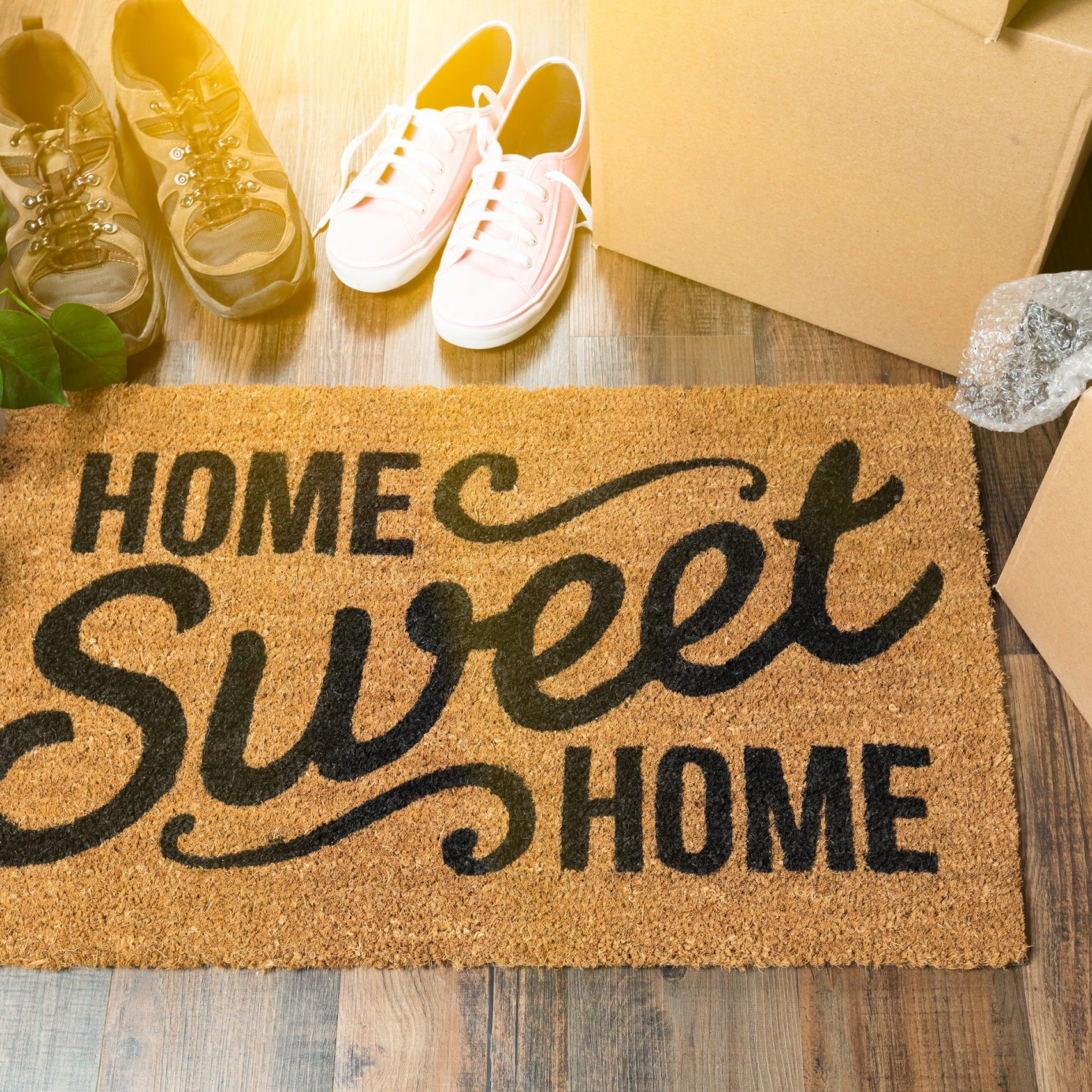 Home Sweet Home Welcome Mat, Moving Boxes, Women and Male Shoes and Plant on Hard Wood Floors.